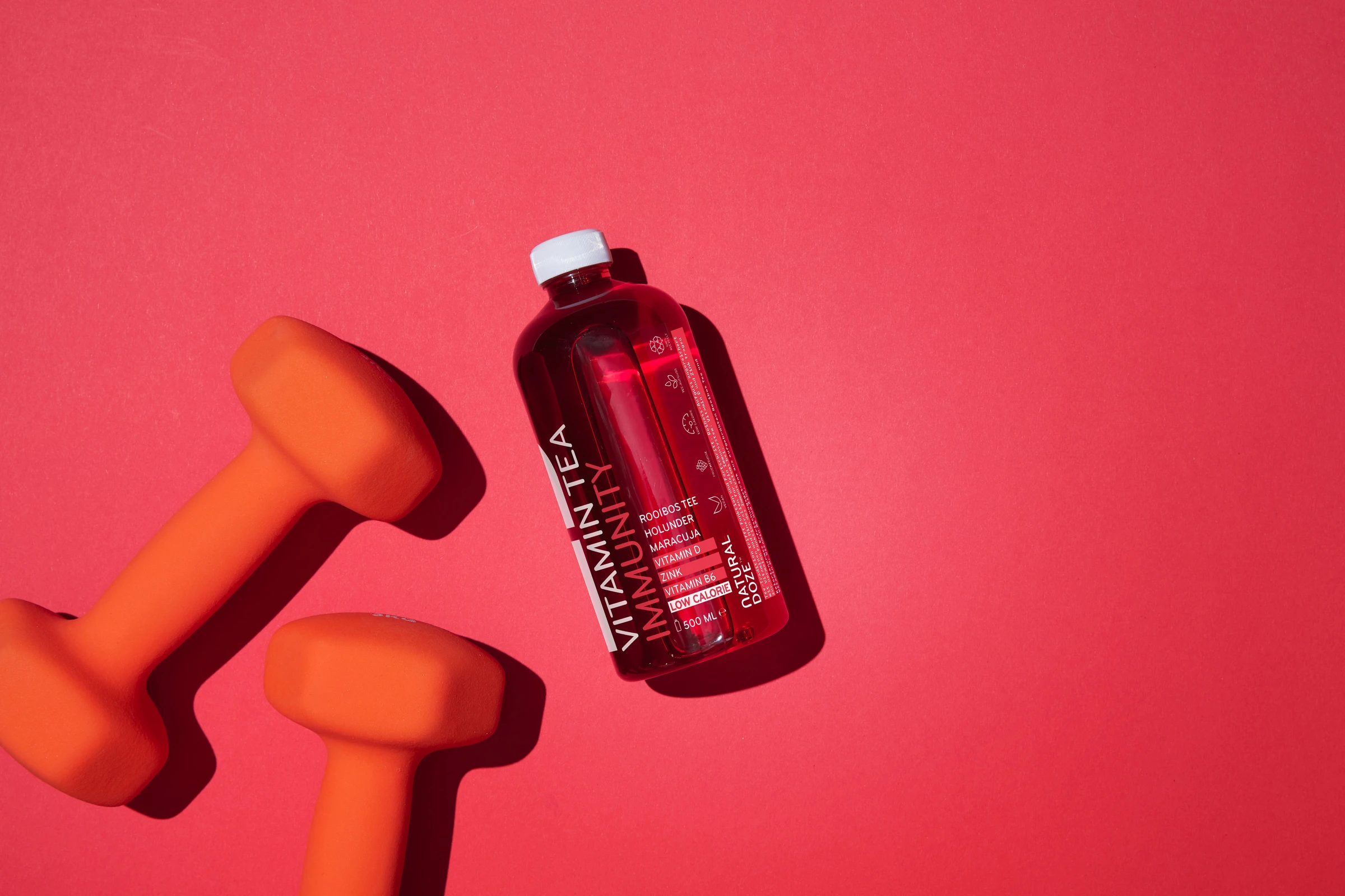 Red Vitamin Tea bottle lying next to orange dumbbells on a red background